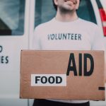 Smiling volunteer holding a box labeled 'Food Aid' outside a van with 'Food Supplies' sign.